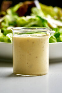 Caesar salad dressing in a glass beaker in front of a plate of lettuce.