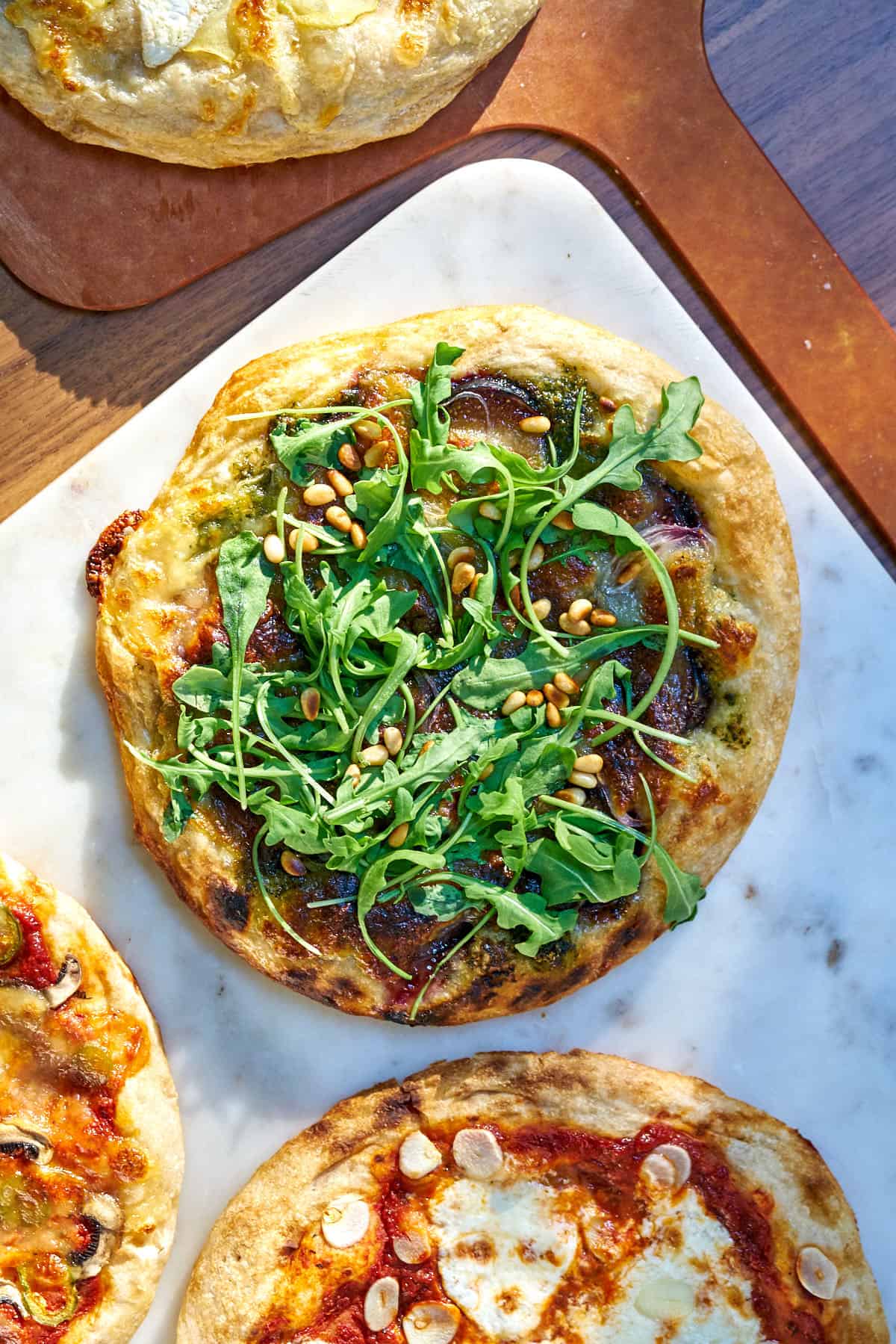 A baked pizza topped with fig and arugula on white marble next to more pizza.