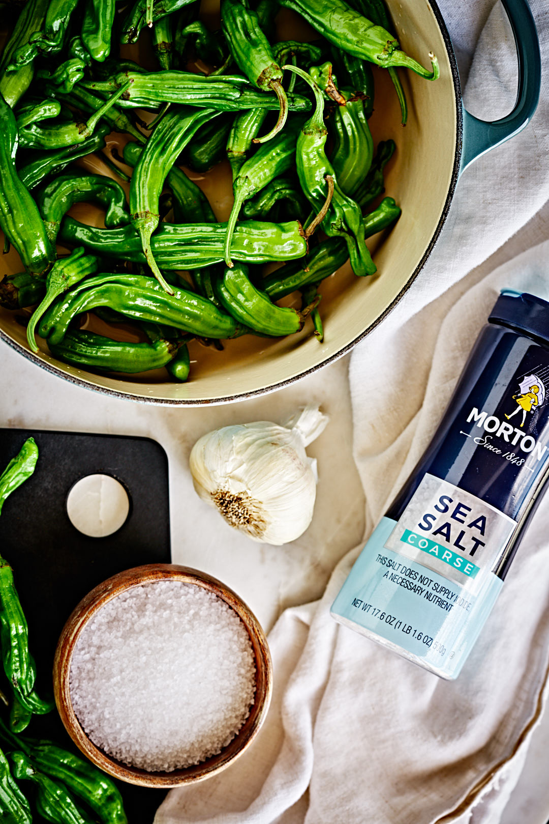 Flatlay of shishito peppers, salt, and a garlic bulb on a tea towel.