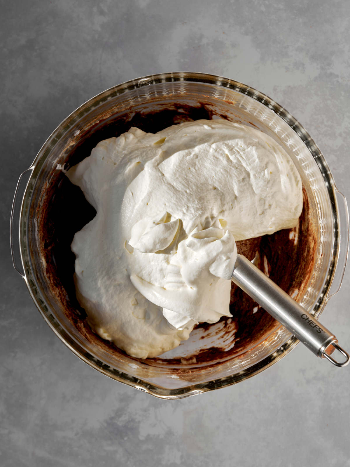 Whipped cream on top of chocolate in a large glass bowl with a metal handle coming out of it.