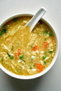 Bowl of yellow egg drop soup with carrots and peas.