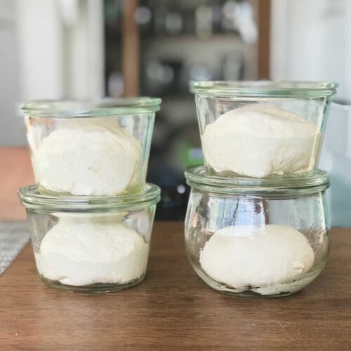 Four glass jars on a wood table with dough in them.