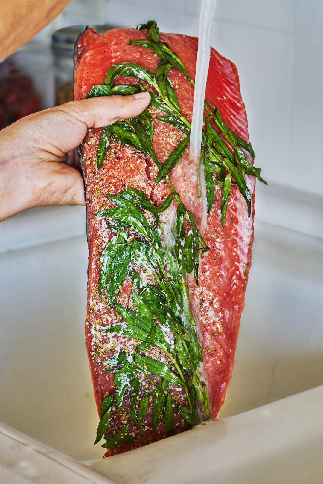 A hand rinsing spices and greens off a large piece of salmon under a faucet.