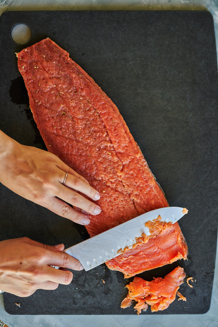 A woman with a silver knife cutting thin slices off a large piece of salmon on a black cutting board.