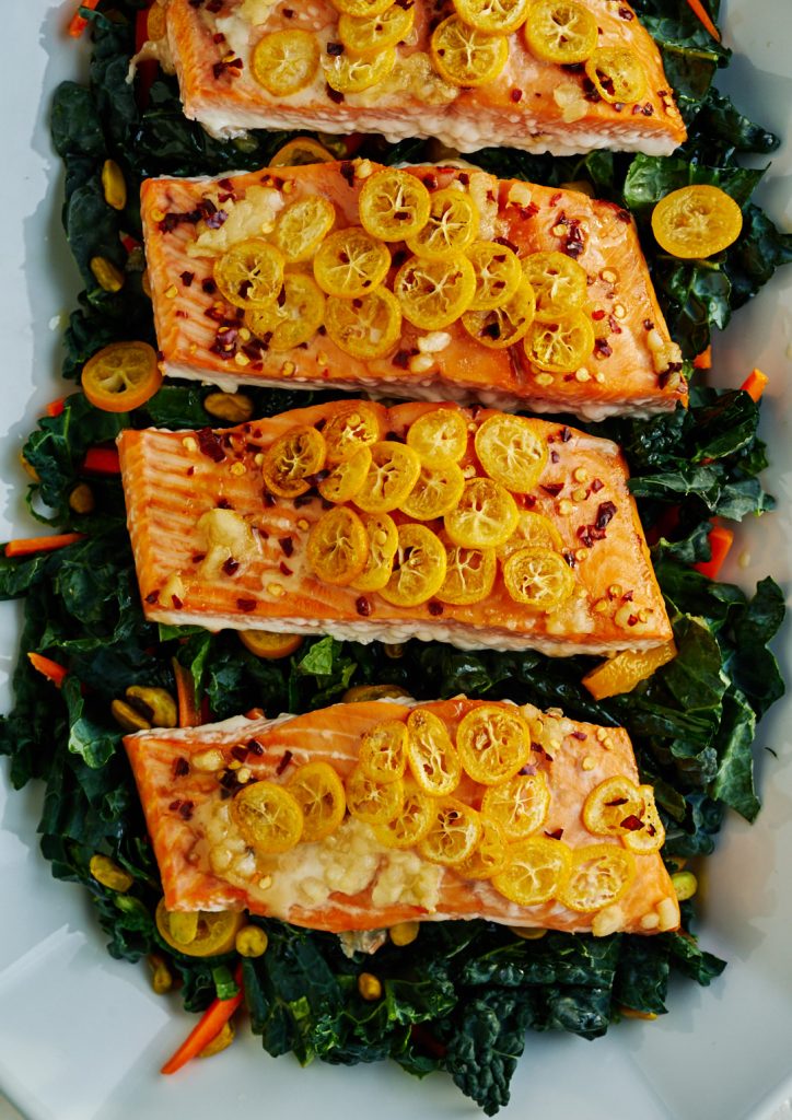 Four salmon steaks covered in slices of yellow kumquat on a bed of greens and kumquats.