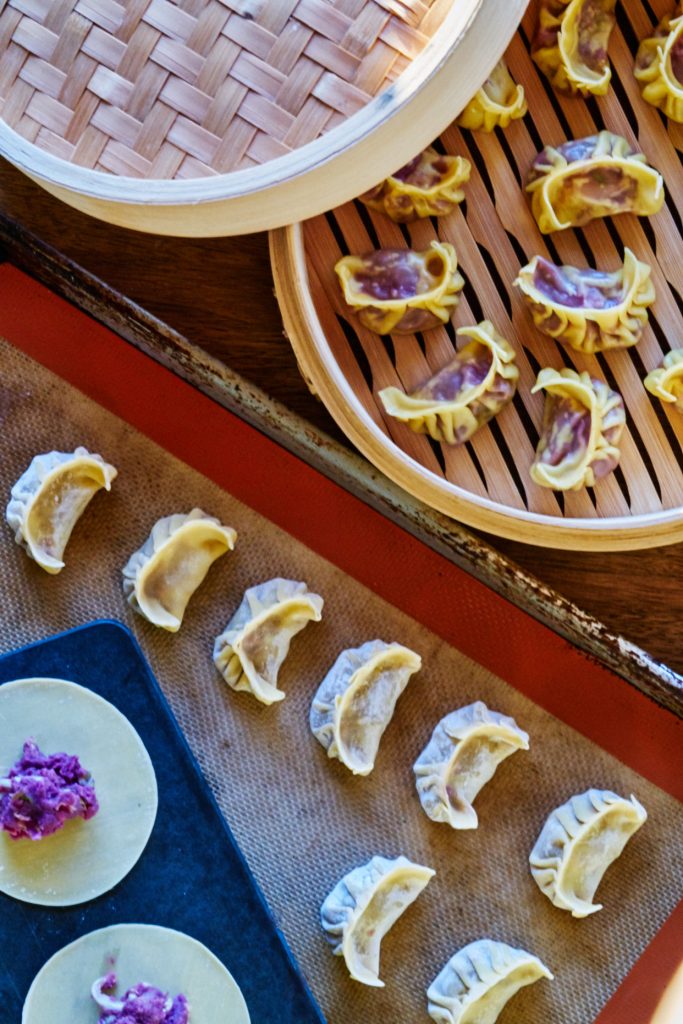 Purple dumplings in a bamboo steamer with uncooked dumplings laid out on a baking sheet below.