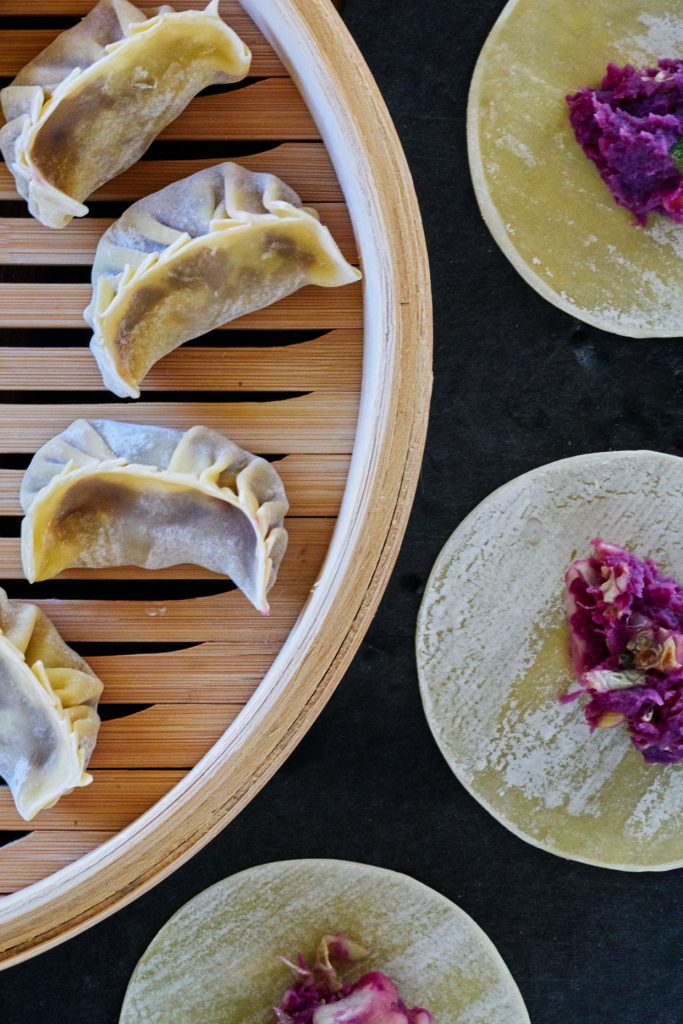 Wrapped purple dumplings in a bamboo steamer with round wonton wrappers and purple filling beside it.