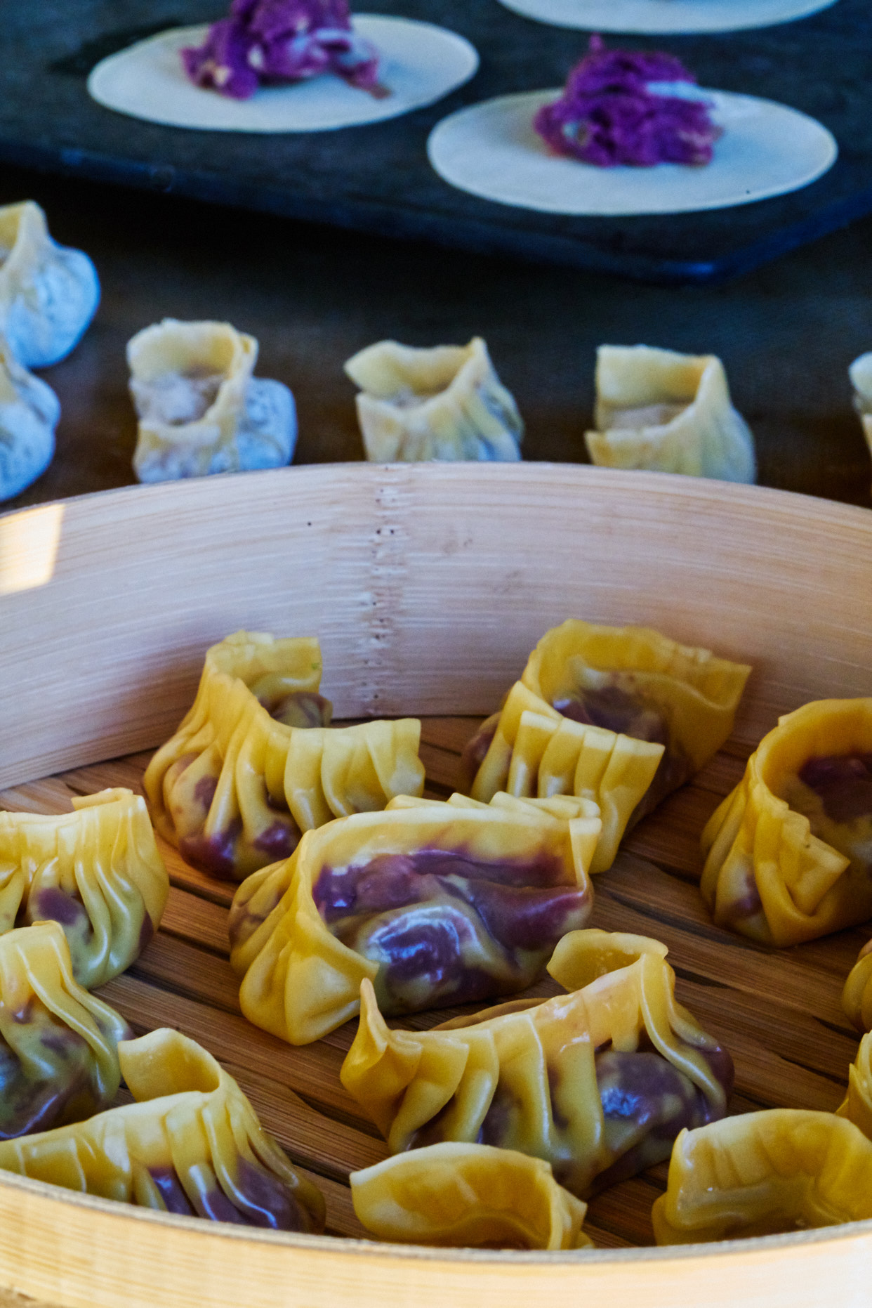 Purple dumplings in a bamboo steamer with uncooked dumplings in the background.