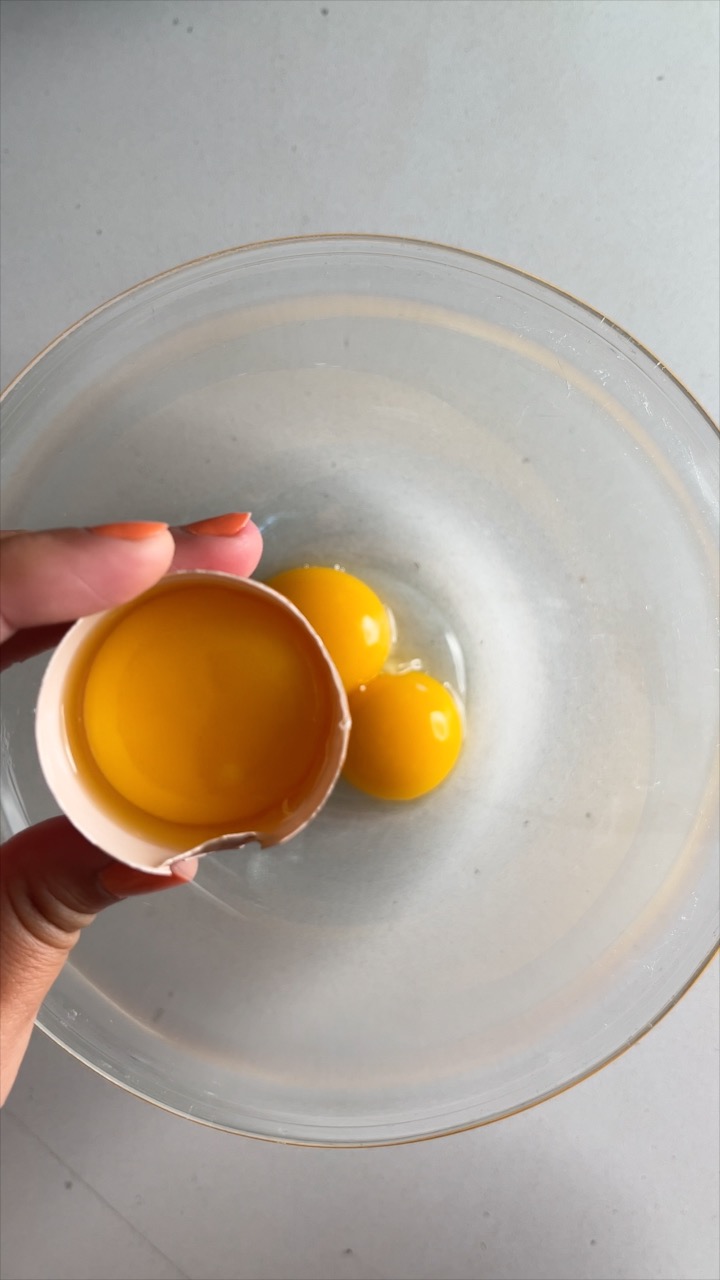 A hand holding an egg yolk in an egg shell over a glass bowl with egg yolks in it.