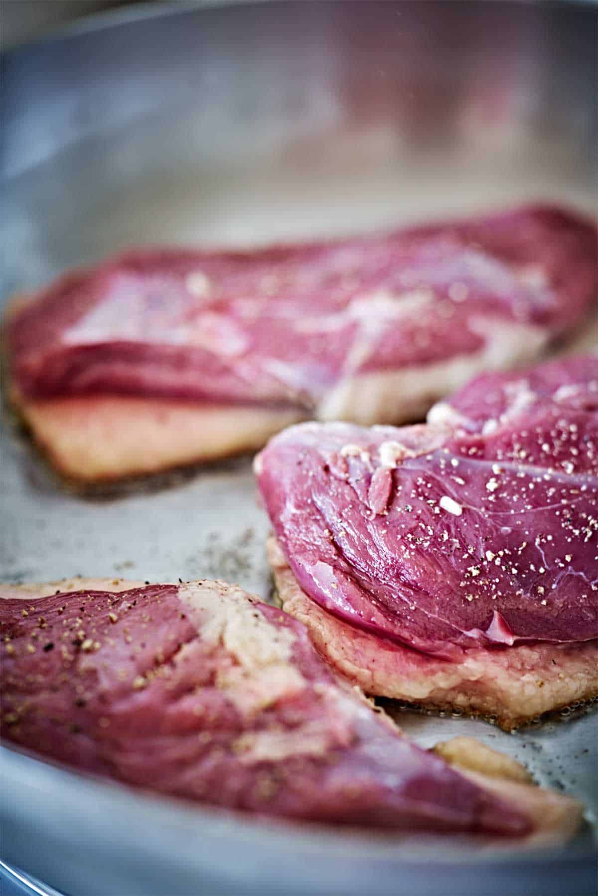 Close up of raw duck breasts cooking in a grey pan.