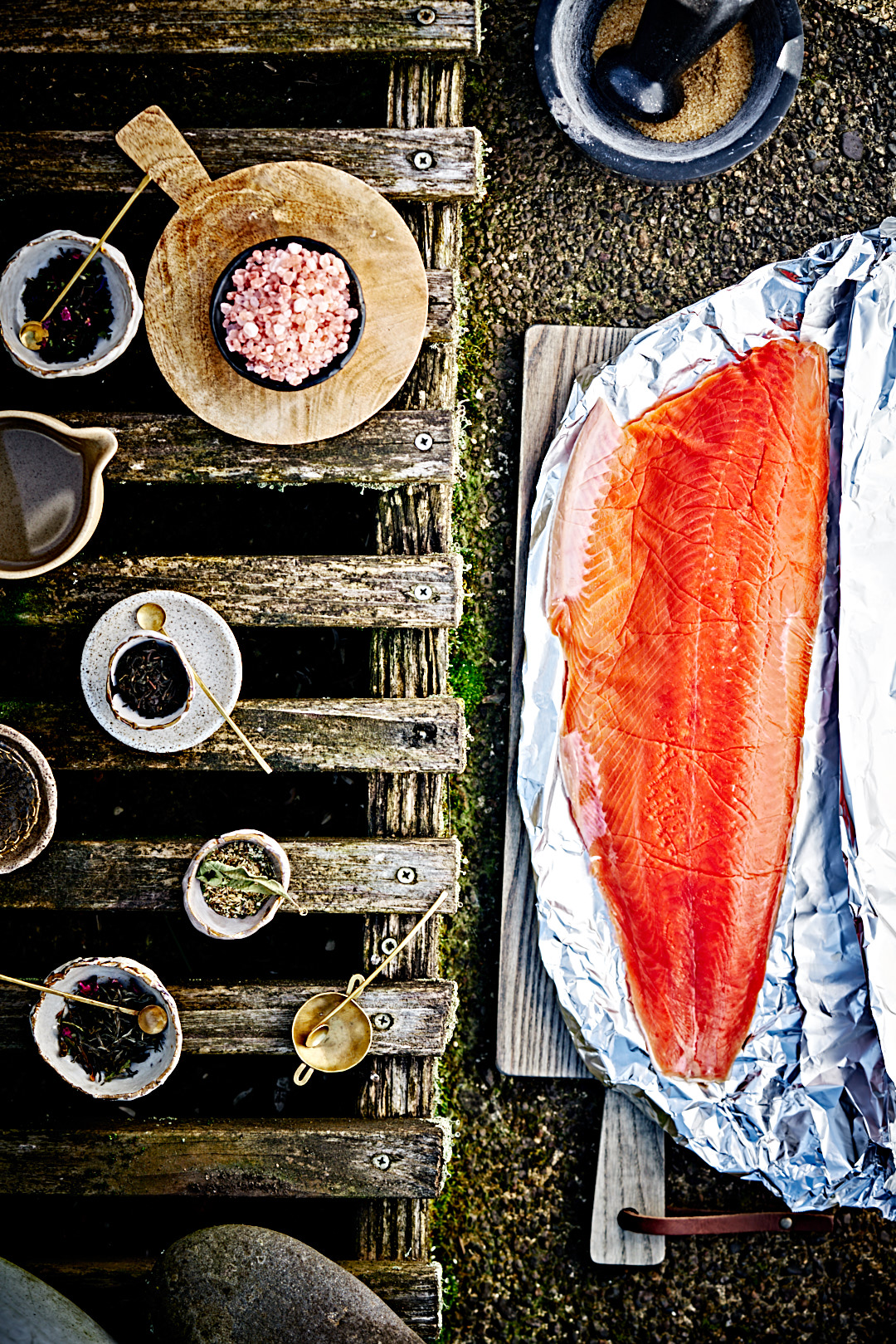 Small bowls of sugar, salt and tea leaves on a wooden bench next to a large fillet of salmon in foil.