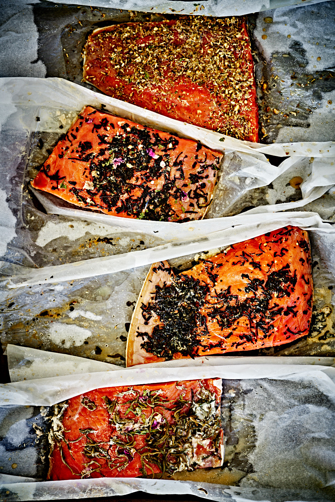 Four cured fillets of salmon covered in sugar, salt, and tea leaves in just-opened parchment paper. 