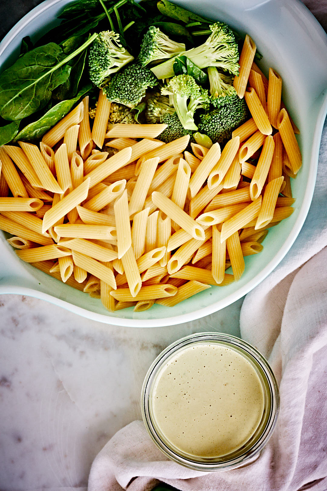 Plain pasta, broccoli and spinach in a large mixing bowl next to a small container of whitish green sauce.