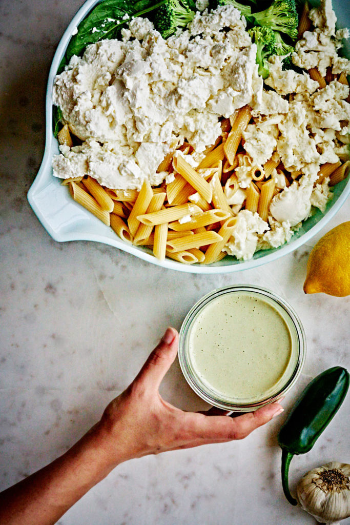 Plain pasta, broccoli, spinach, and mozzarella in a large mixing bowl next to a small container of whitish green sauce.