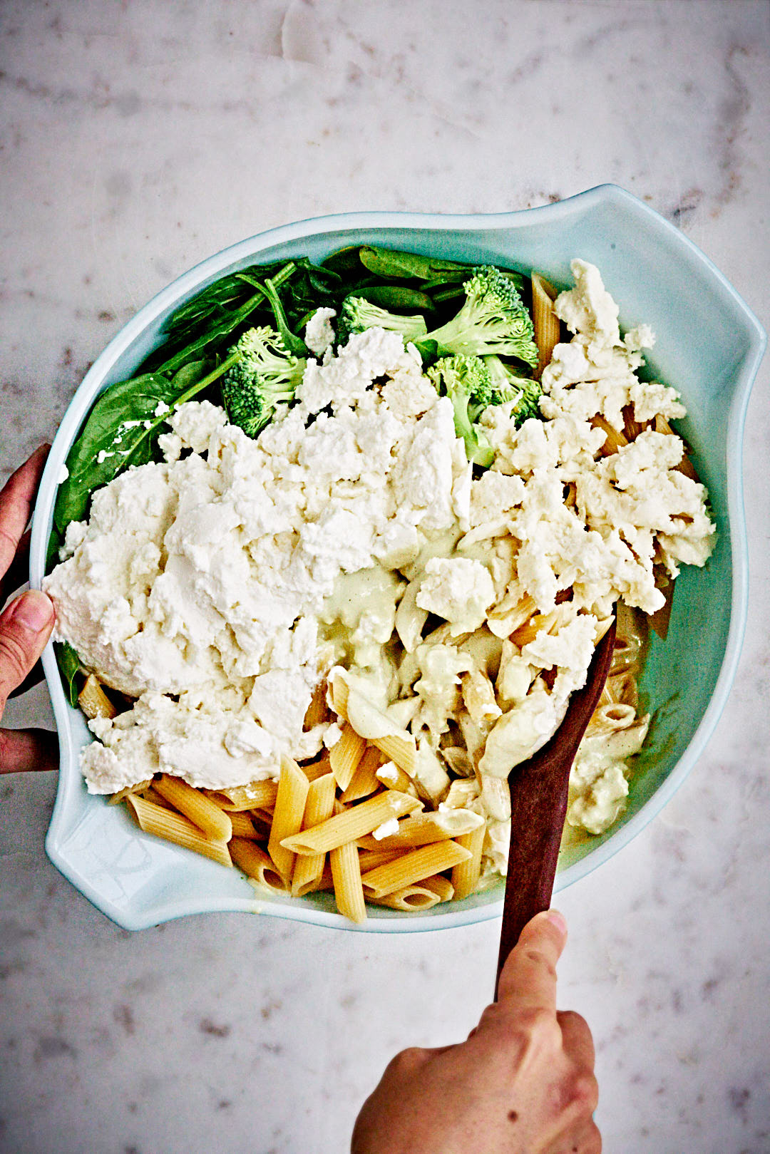 A hand with a wooden spoon mixing large bowl filled with pasta, spinach, broccoli, and mozzarella.