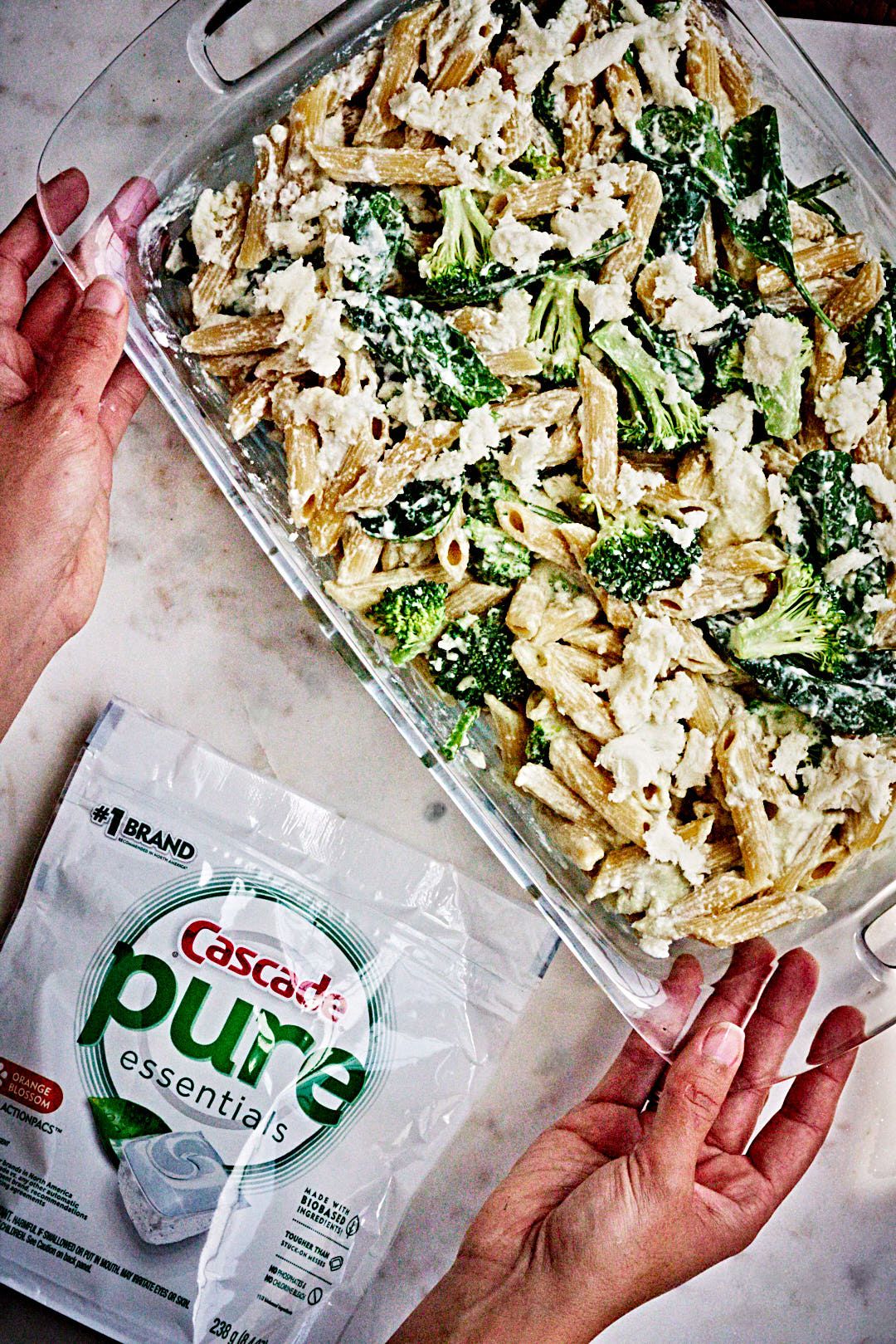 Two hands holding a large glass baking dish filled with pasta, broccoli, spinach, pieces of mozzarella, and a packet of dishwasher tabs next to it.