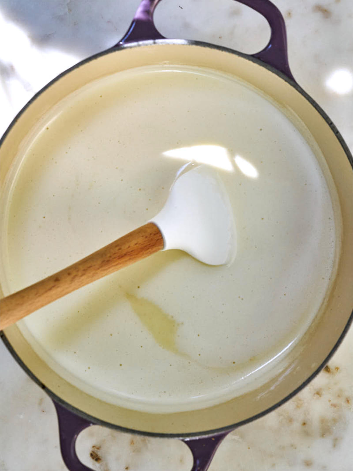 Foamy liquid in a purple enameled pot with a white rubber spatula with a wooden handle.