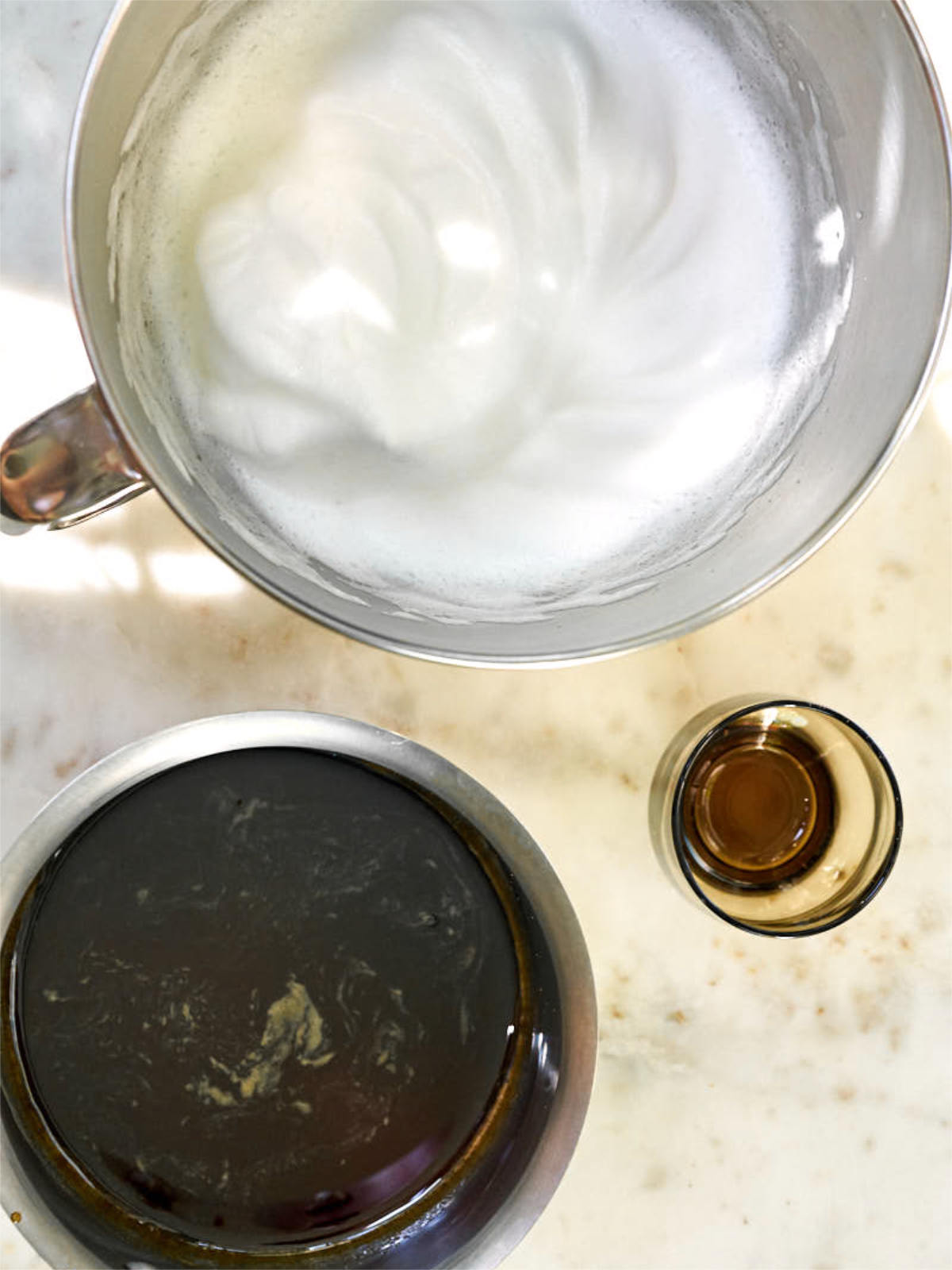 Whipped meringue in a stand mixer bowl next to dark brown simple syrup and a brown glass.