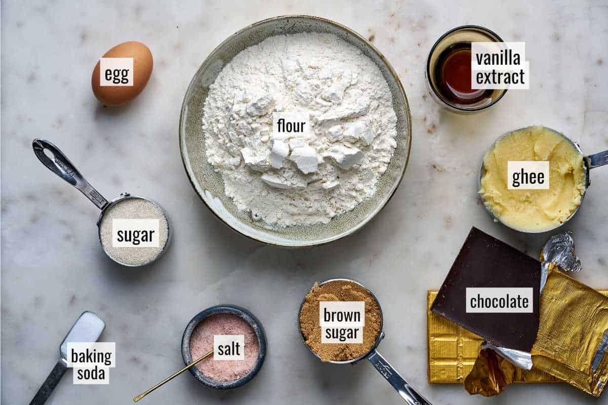 Ingredients for chocolate chip cookies with text labels on a marble countertop.