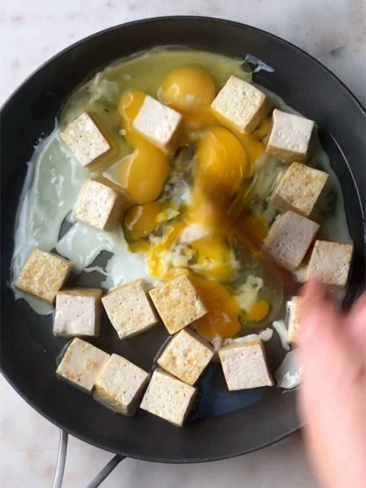 A hand using a wooden spoon to scramble eggs in a pan with tofu cubes.