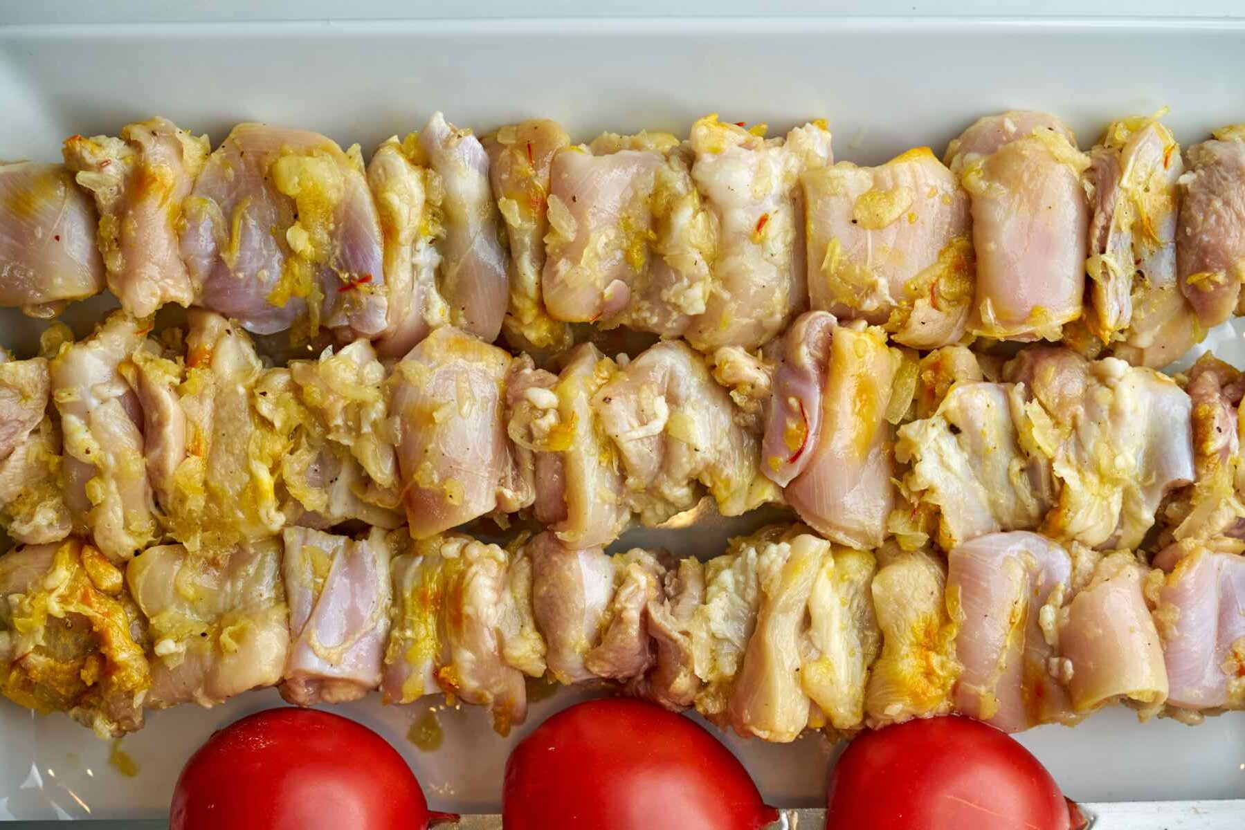 Raw chicken kabobs on a platter next to tomatoes.