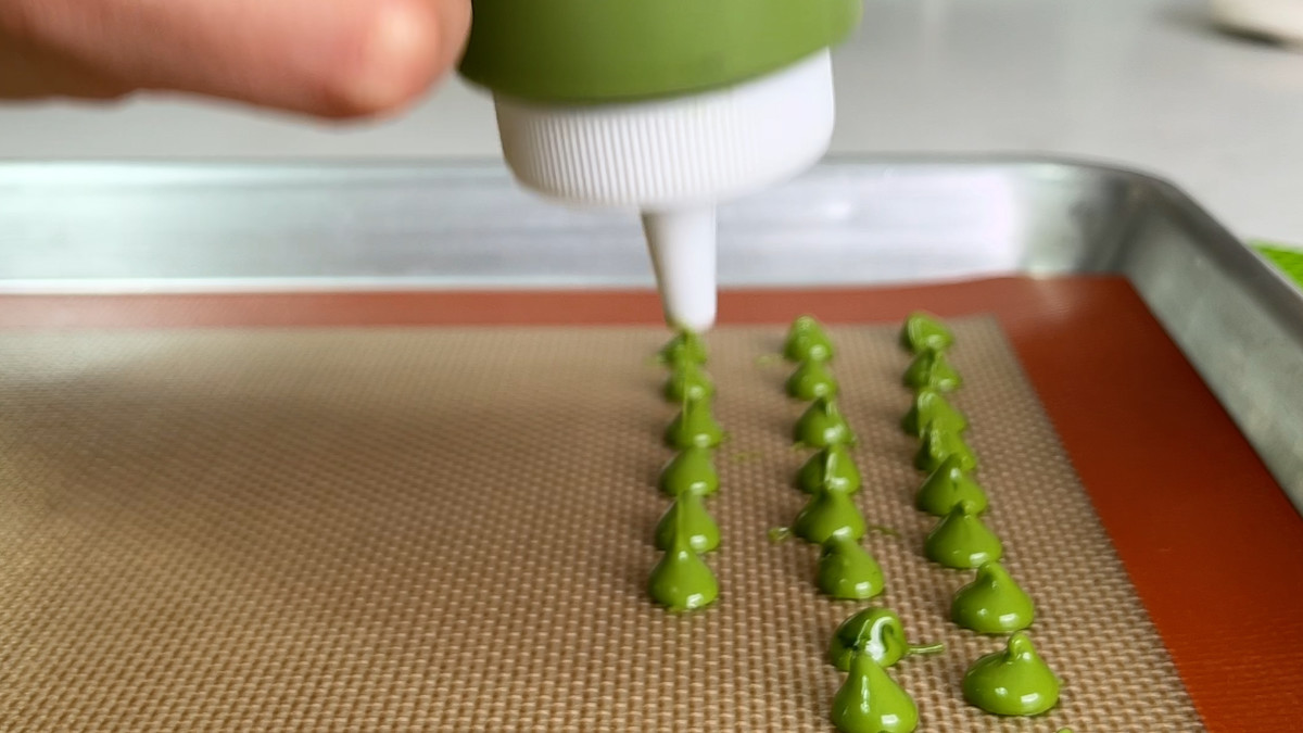 A hand piping green chocolate chips with a squeeze bottle on a baking mat.