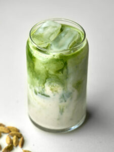 Side view of a glass soda can with an iced green match latte next to cardamom pods.