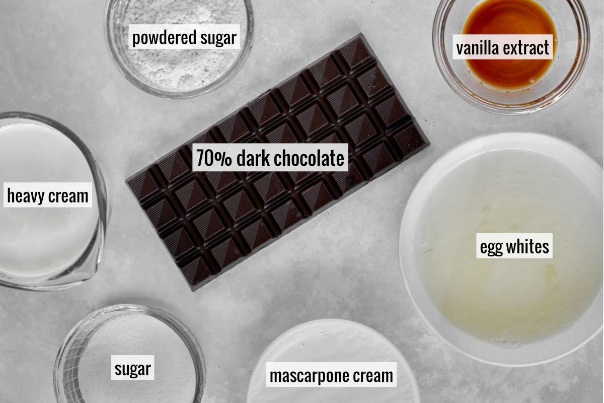 A large bar of chocolate next to bowls with other ingredients like cream, egg whites, vanilla extract, and sugar.