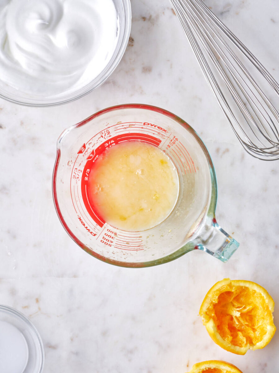 Orange juice in a liquid measuring cup next to an orange half, whisk, and bowl with whipped egg whites.