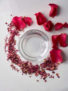 Dried and fresh rose petals surrounding a glass bowl filled with clear rose water.