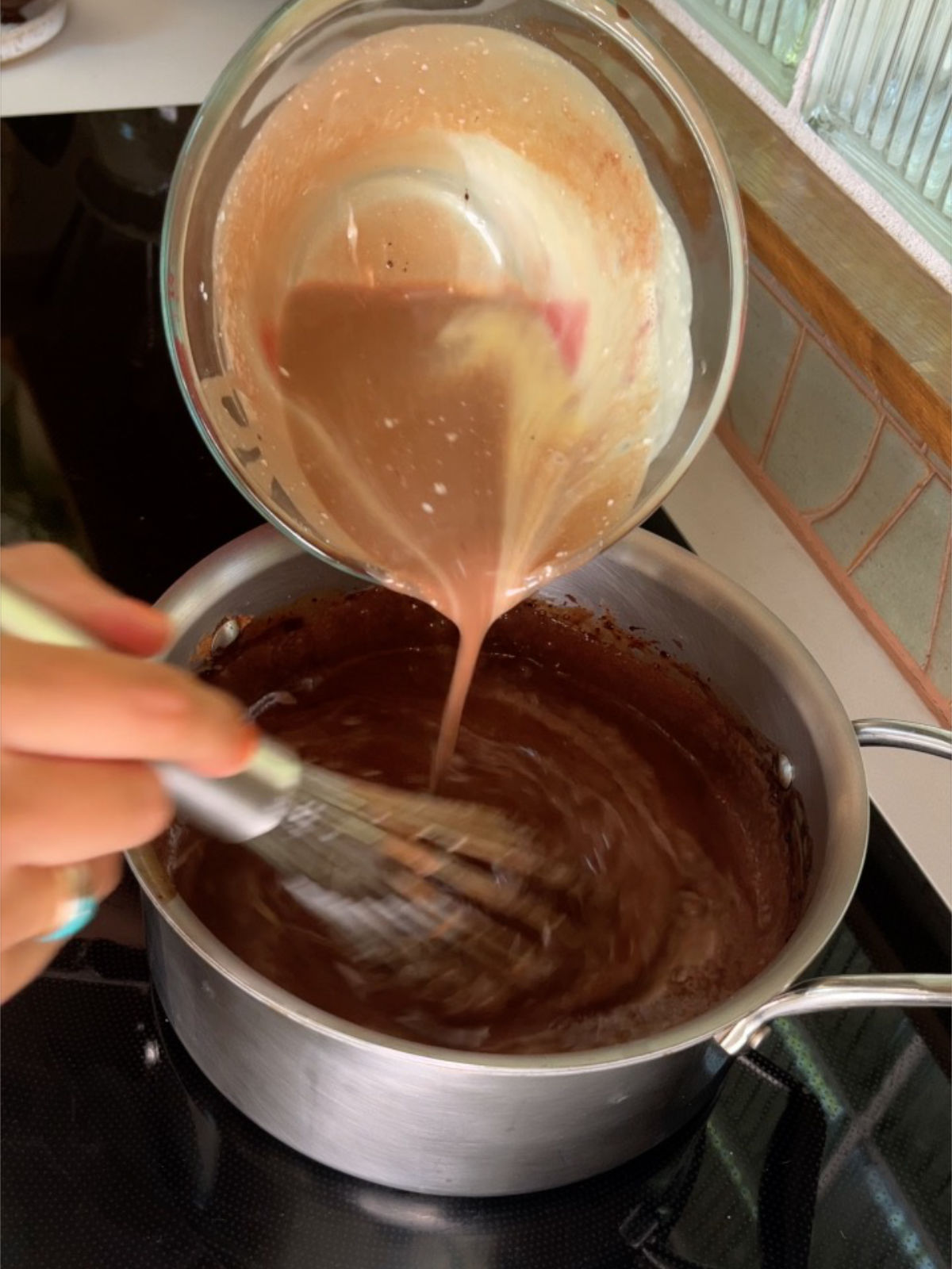 Pouring light brown liquid from a liquid measuring cup into a metal pot with chocolate liquid and a hand whisking the pot contents.