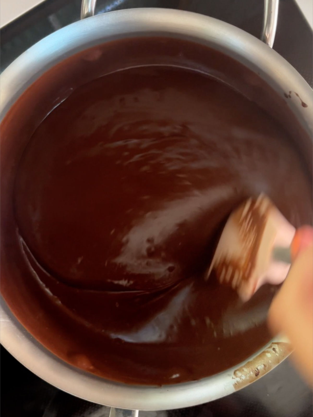 Pink rubber spatula stirring thickened chocolate pudding in a metal pot.