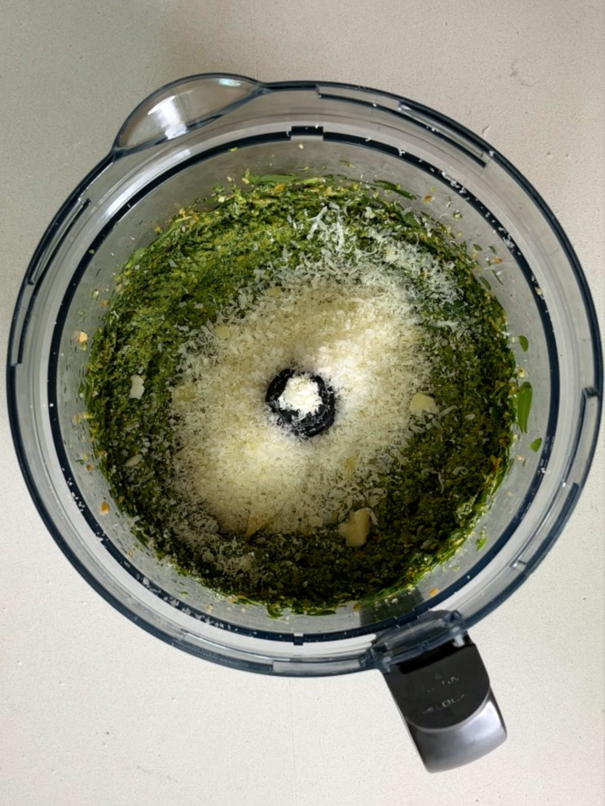 Pesto in the bowl of a food processor with a topping of grated parmesan.