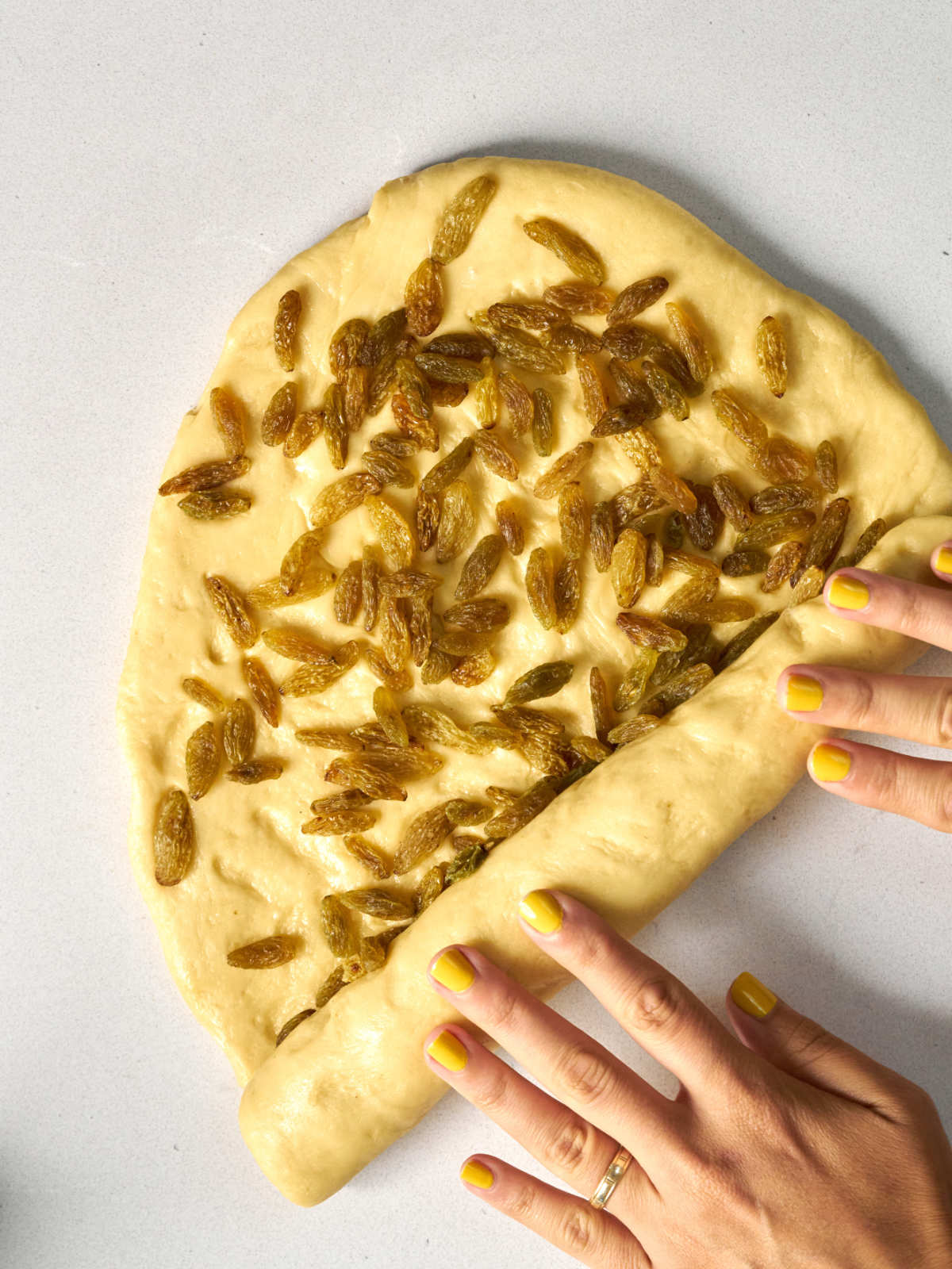 Two hands with yellow nail polish rolling dough covered in an even layer of green raisins.