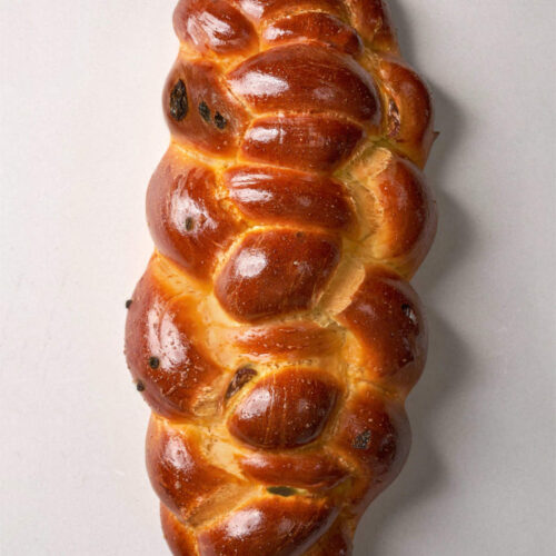 A brown, baked, braided challah bread with raisins on a light grey background.