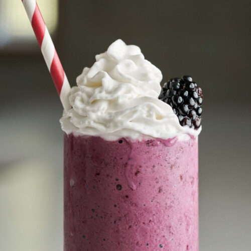 Purple milkshake topped with whipped cream, a blackberry, and a striped straw.