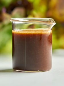 Salad dressing in a small beaker in front of lettuce.