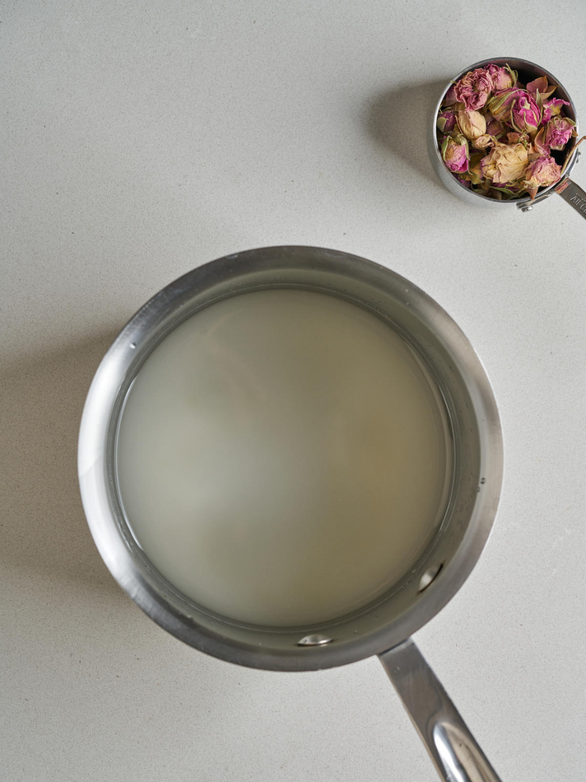 A saucepan with sugar and water next to dried rose petals.