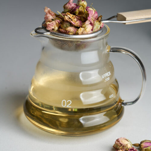 A glass pitcher with yellow simple syrup next to dried rose petals and with a strainer filled with dried roses on top of the pitcher.