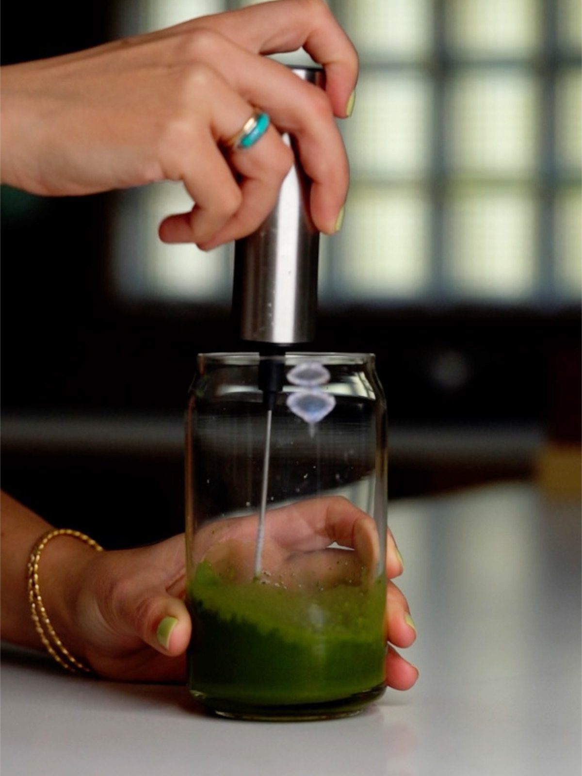 Hands with green nails and a turquoise ring frothing matcha in a tall glass on a countertop with a handheld frother.