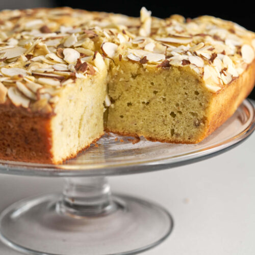 A beige square cake with an almond topping sitting on a glass cake display stand.