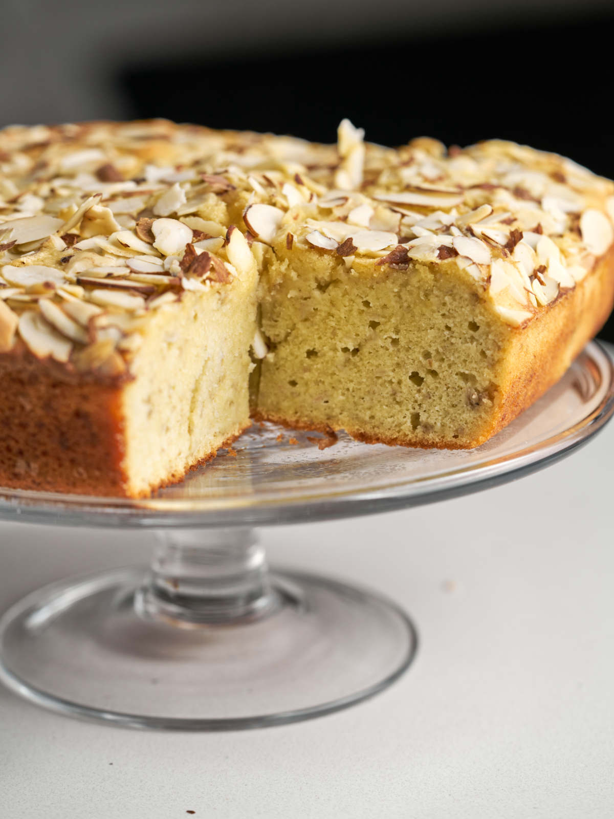 A beige square cake with an almond topping sitting on a glass cake display stand.