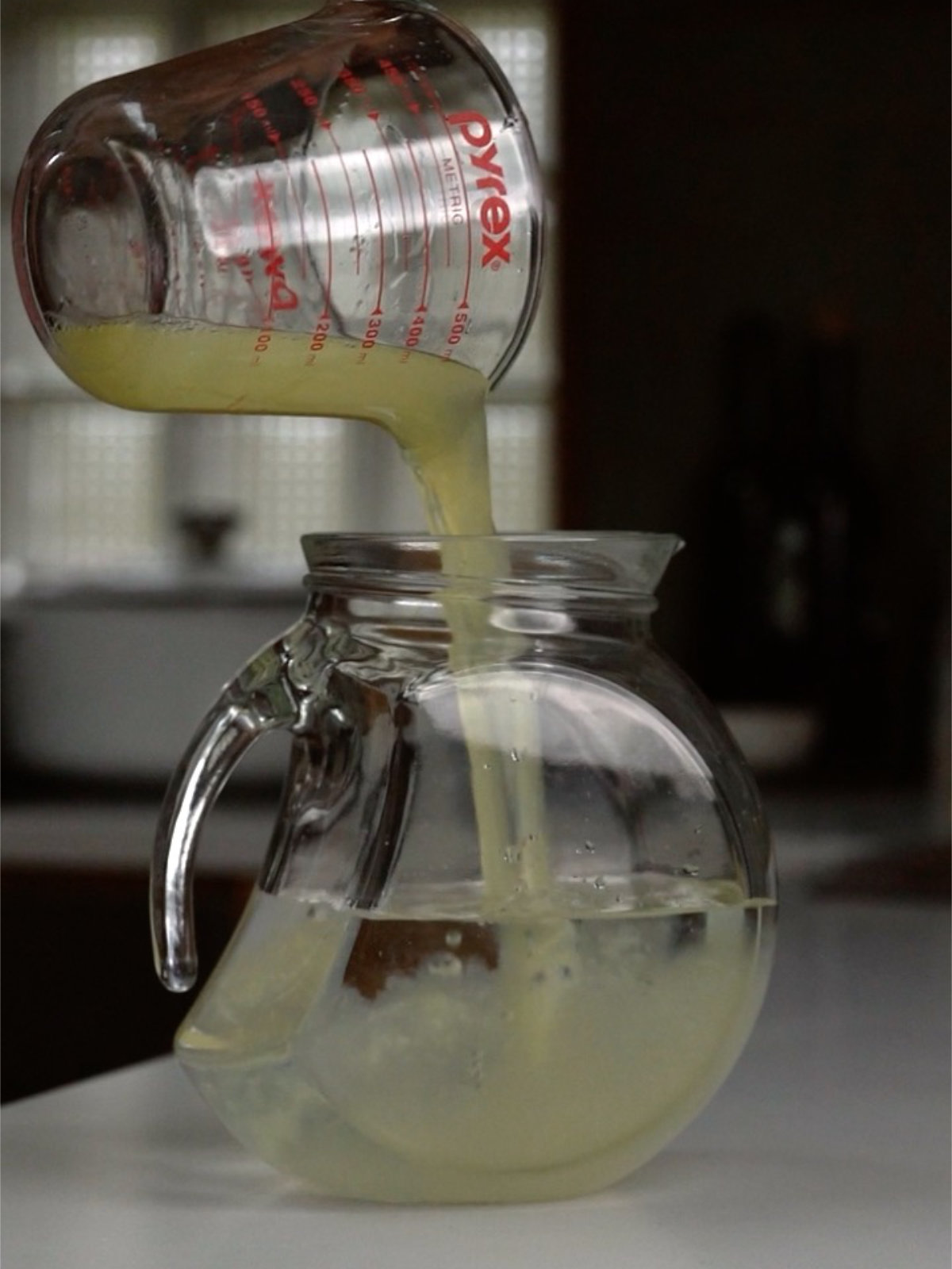 Pouring lemon juice into a glass pitcher from a liquid measuring cup on a countertop.