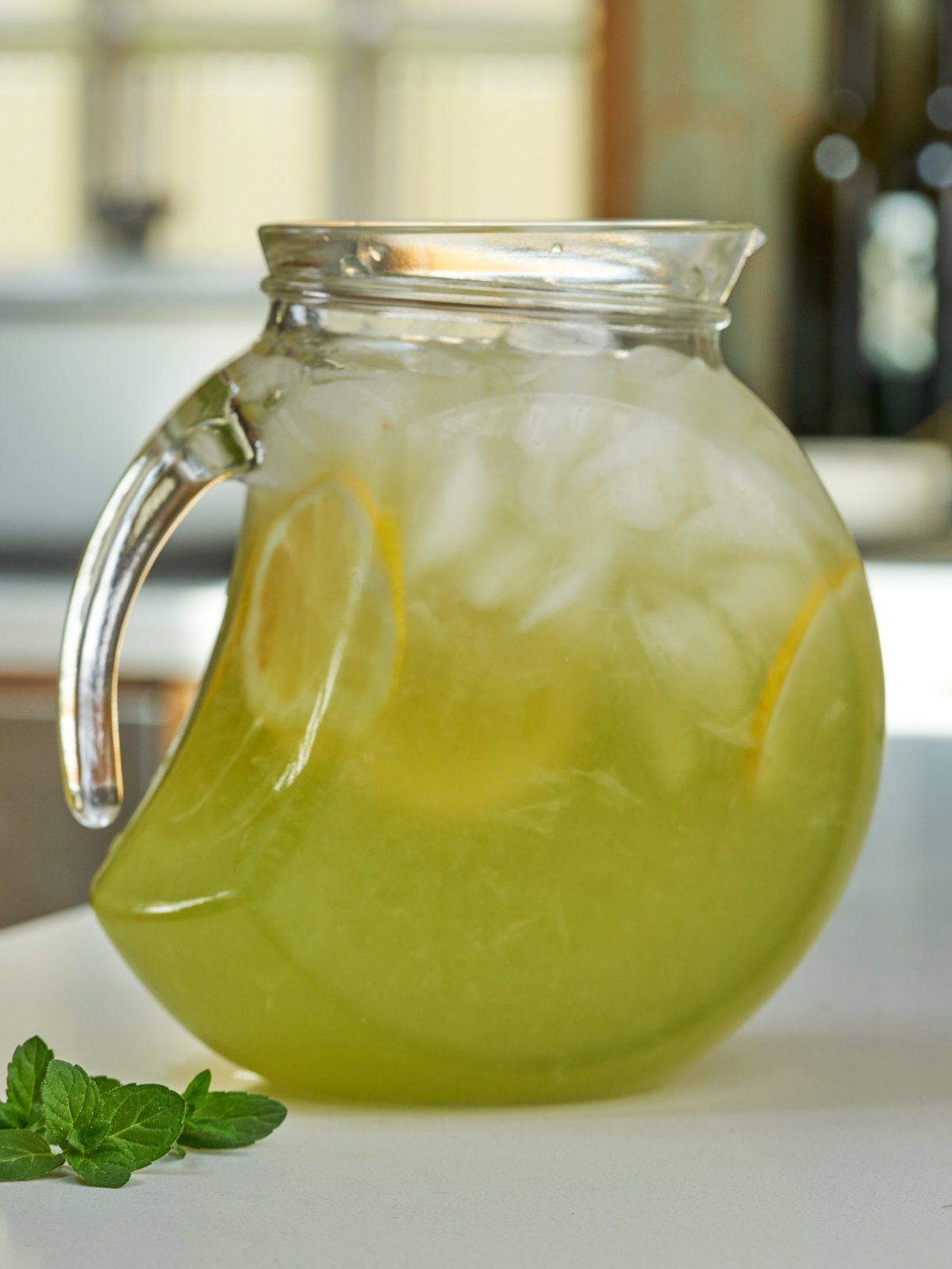 Pitcher with green lemonade, lemon slices, and ice next to mint leaves.