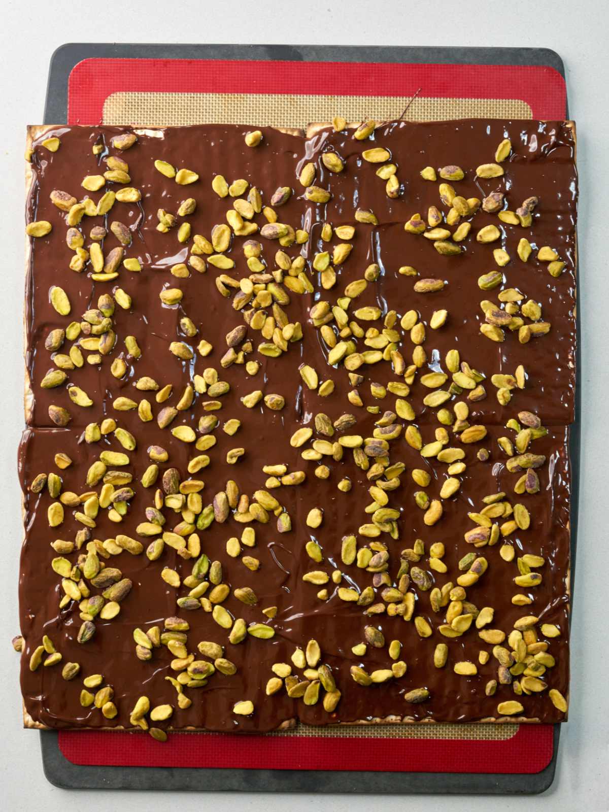 A large square matzo covered in melted chocolate and pistachios on top of a baking mat.