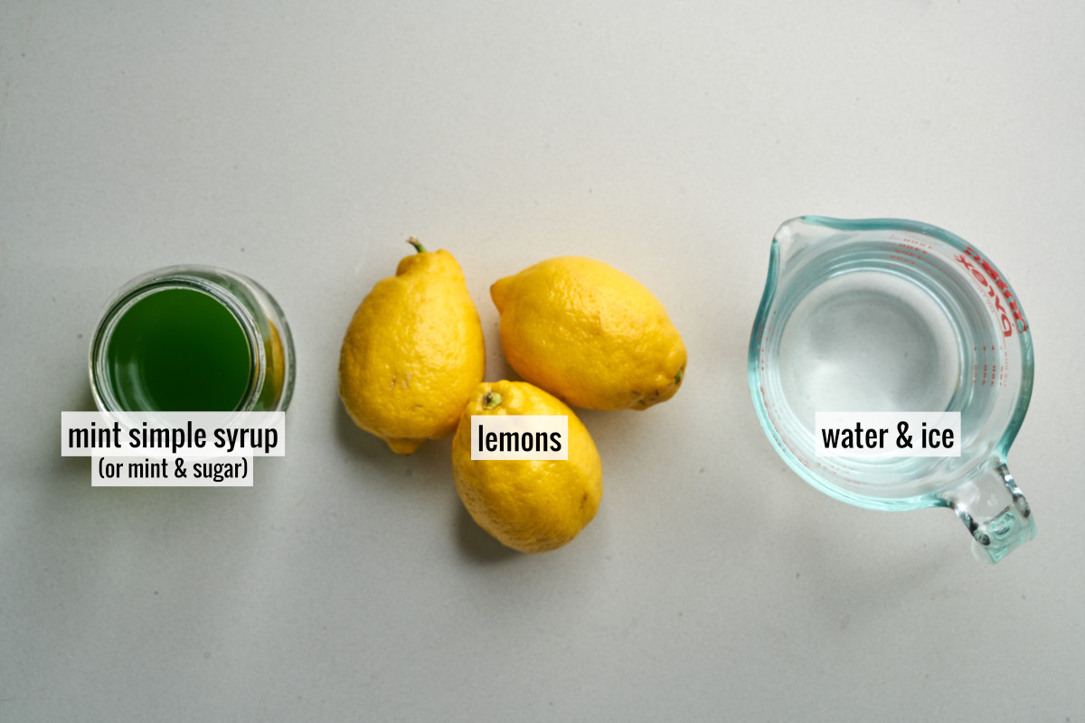 Three lemons next to a liquid measuring cup with water and a small jar of bright green simple syrup.