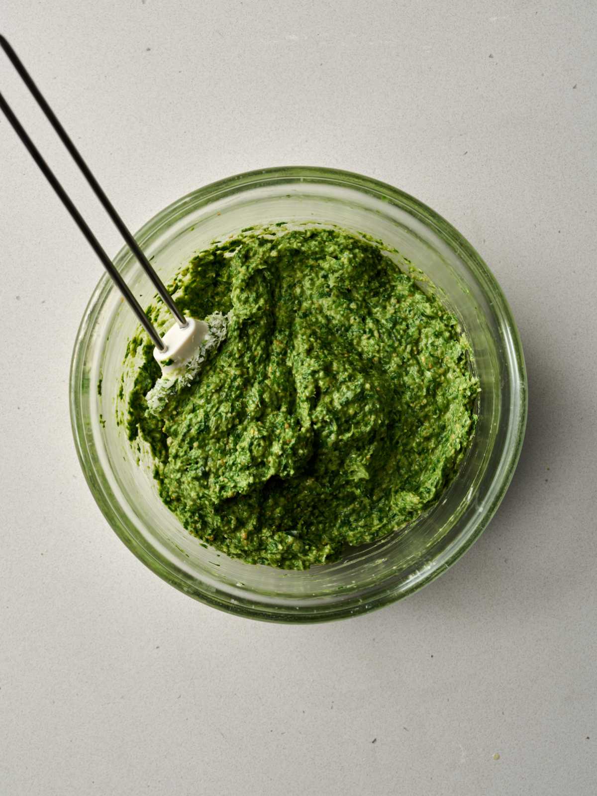 White spatula stirring green pesto in a large glass bowl.