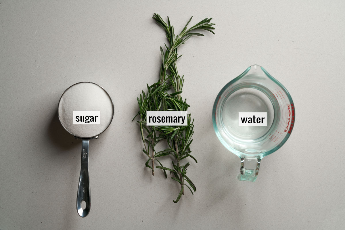 Photograph of three ingredients including sugar rosemary and water in a glass measuring jug