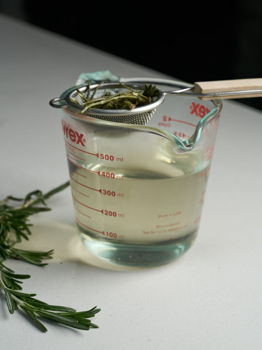 A glass measuring cup filled with simple syrup and a strainer balanced on top filled with rosemary leaves.