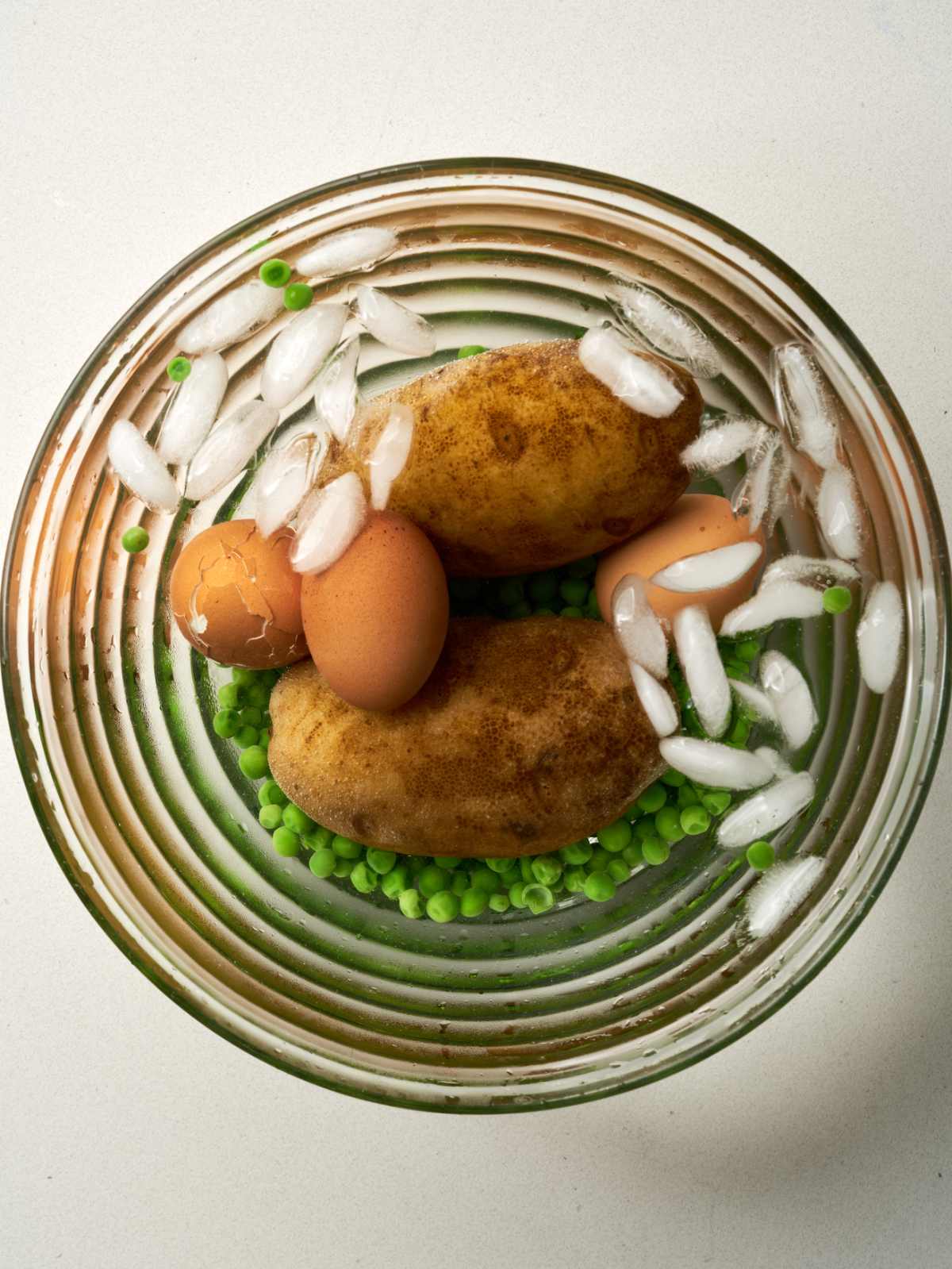 Potatoes, eggs, and green peas in a glass bowl with ice water.